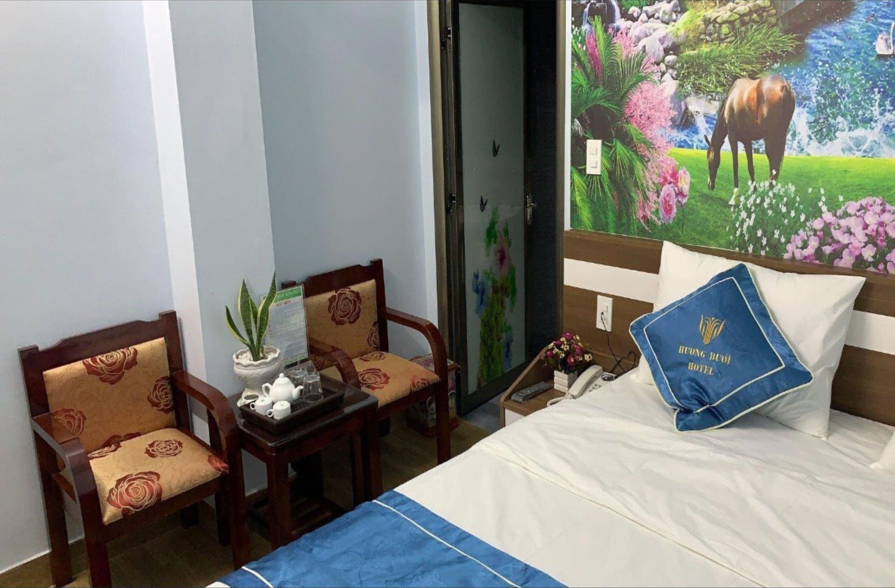 Une chambre d’hôtel avec un lit, deux chaises, une petite table avec une théière et une fresque murale.