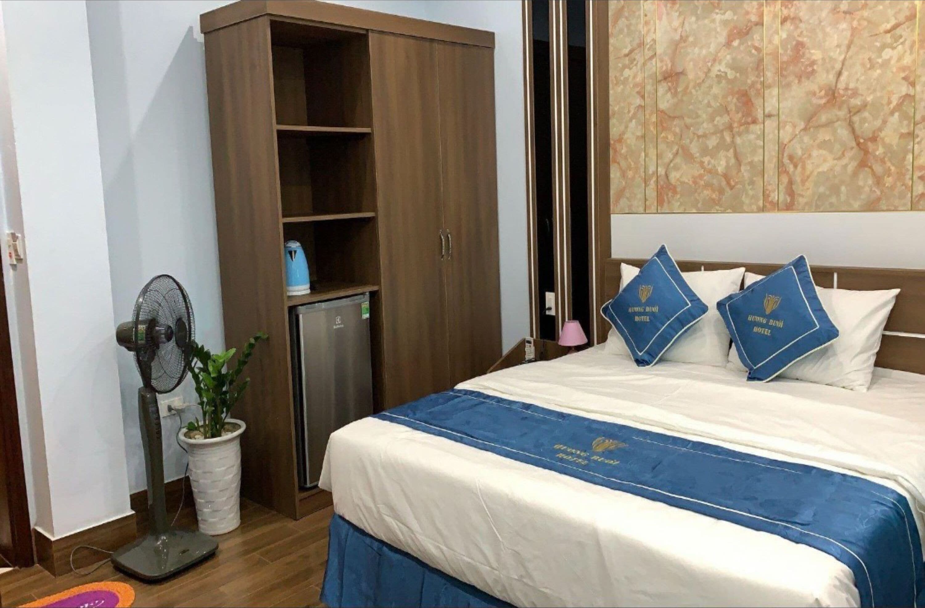 Une chambre d’hôtel avec un lit, une armoire, un ventilateur, une plante en pot et un mur décoratif.
