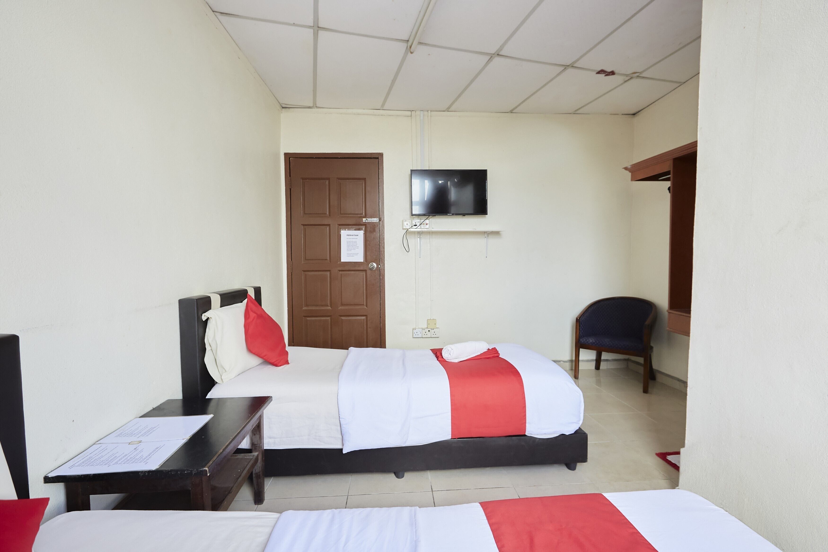 Ein Hotelzimmer mit zwei Betten, einem an der Wand montierten Fernseher, einer Tür und einem Sessel.
