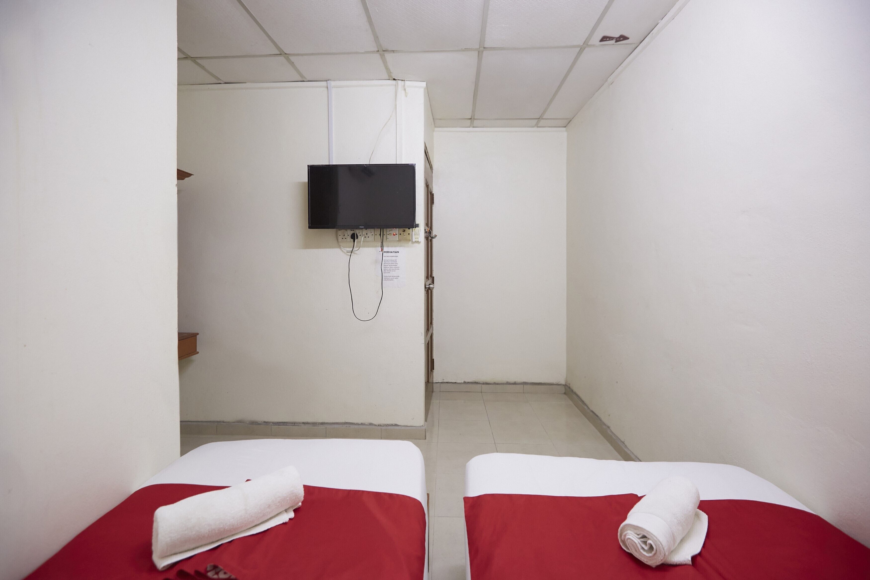 A small, narrow room with two beds, each with a red bedspread and a white towel. A television is mounted on the wall, and there is a door at the end of the room.