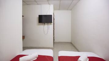 A small, narrow room with two beds, each with a red bedspread and a white towel. A television is mounted on the wall, and there is a door at the end of the room.