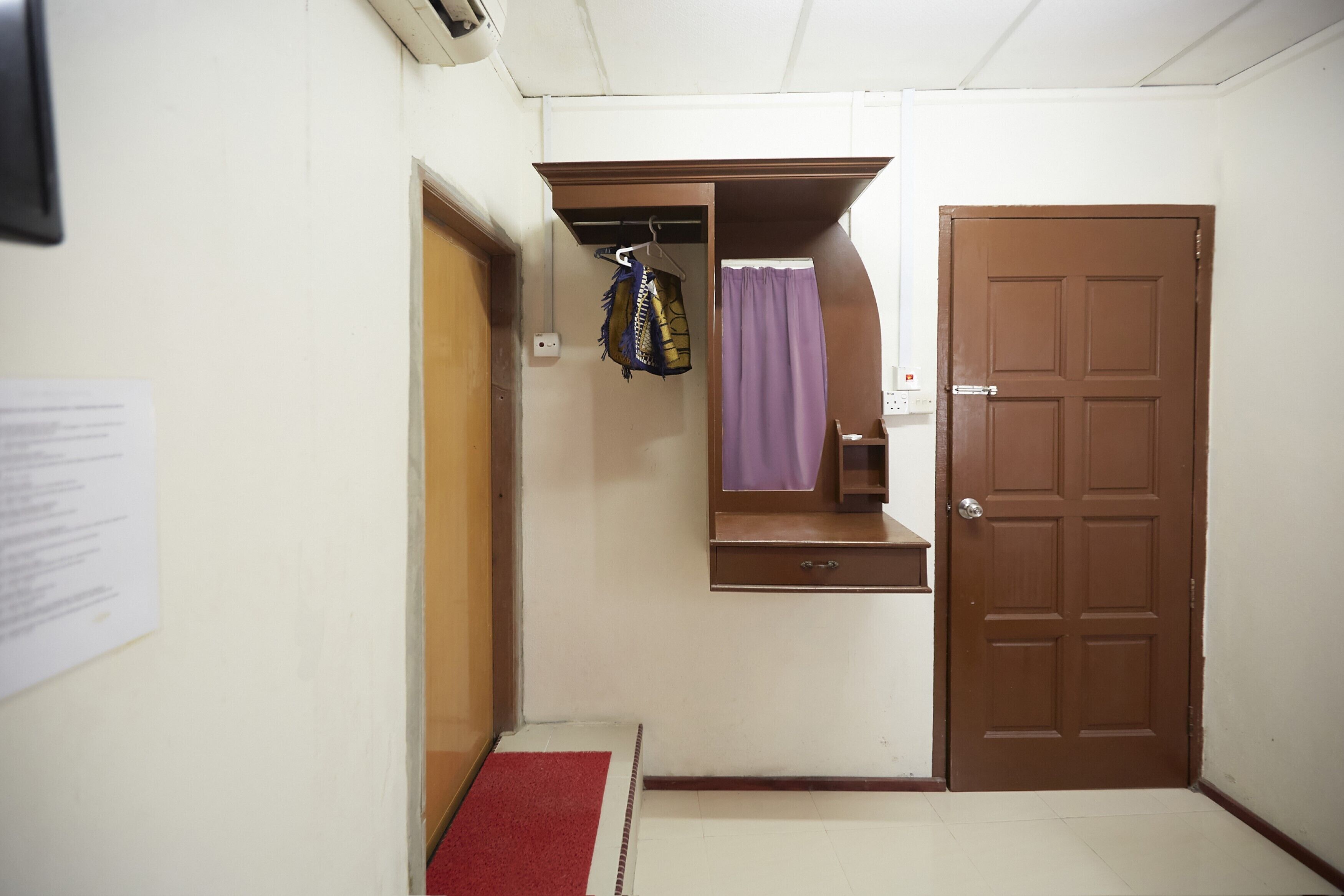 A small room with a brown door, a red carpet, a hanging garment, and a wall-mounted shelf.