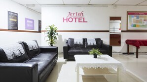 A hotel reception area with two black leather sofas, a white table with a lace tablecloth, and a potted plant. The reception desk has a sign reading "Jerteh HOTEL" and motivational posters on the wall. - Jerteh Hotel, Terengganu (Jerteh)