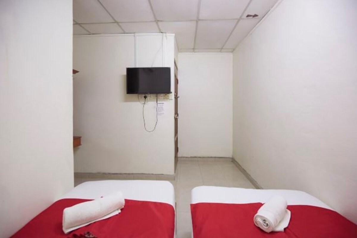 Une petite pièce étroite, avec deux lits, chacun recouvert d’une couverture rouge et agrémenté d’oreillers blancs. Un téléviseur est fixé au mur, au-dessus d’une porte.