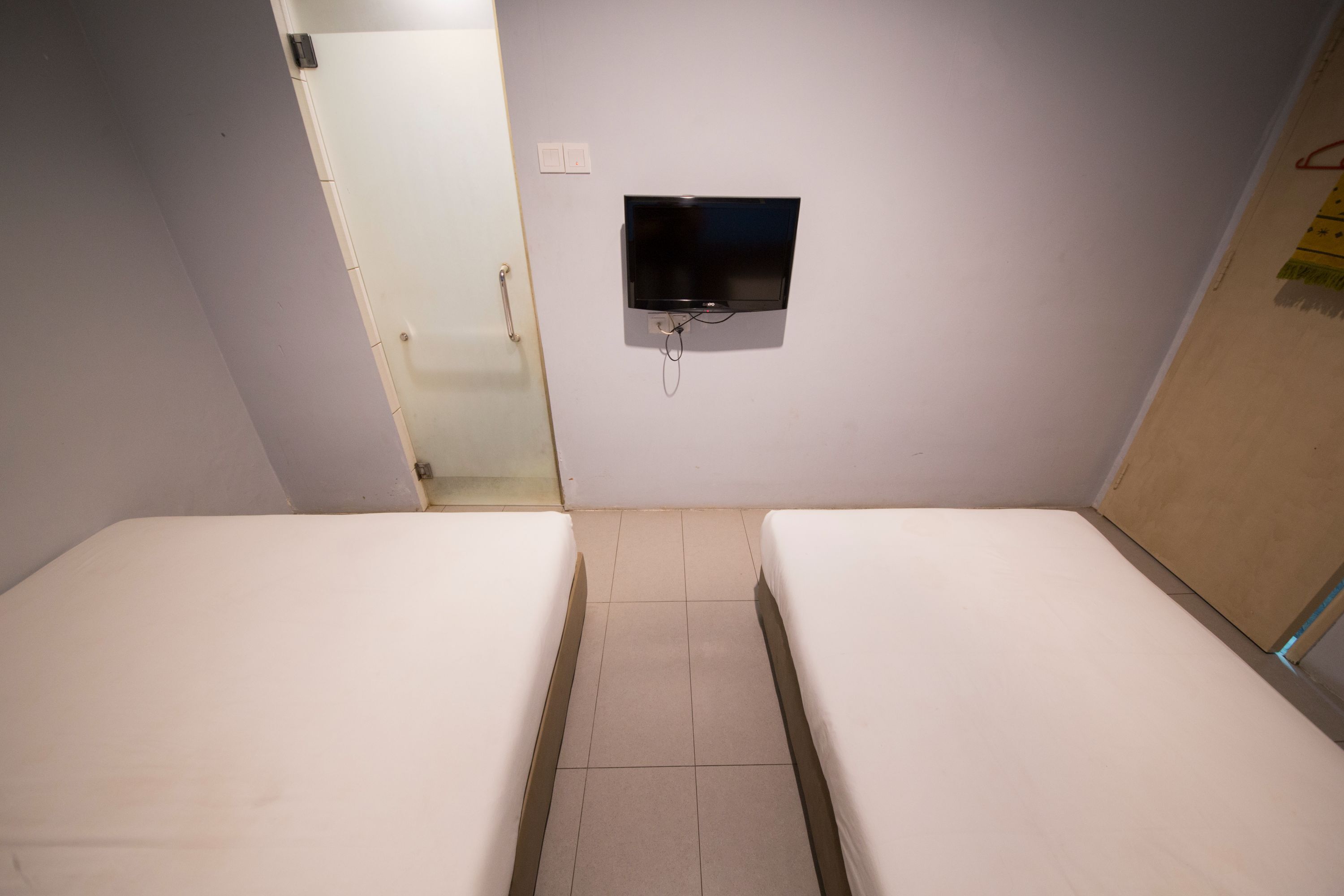 Una habitación pequeña y sencilla con dos camas, un televisor montado en la pared y una puerta que da a otra habitación.
