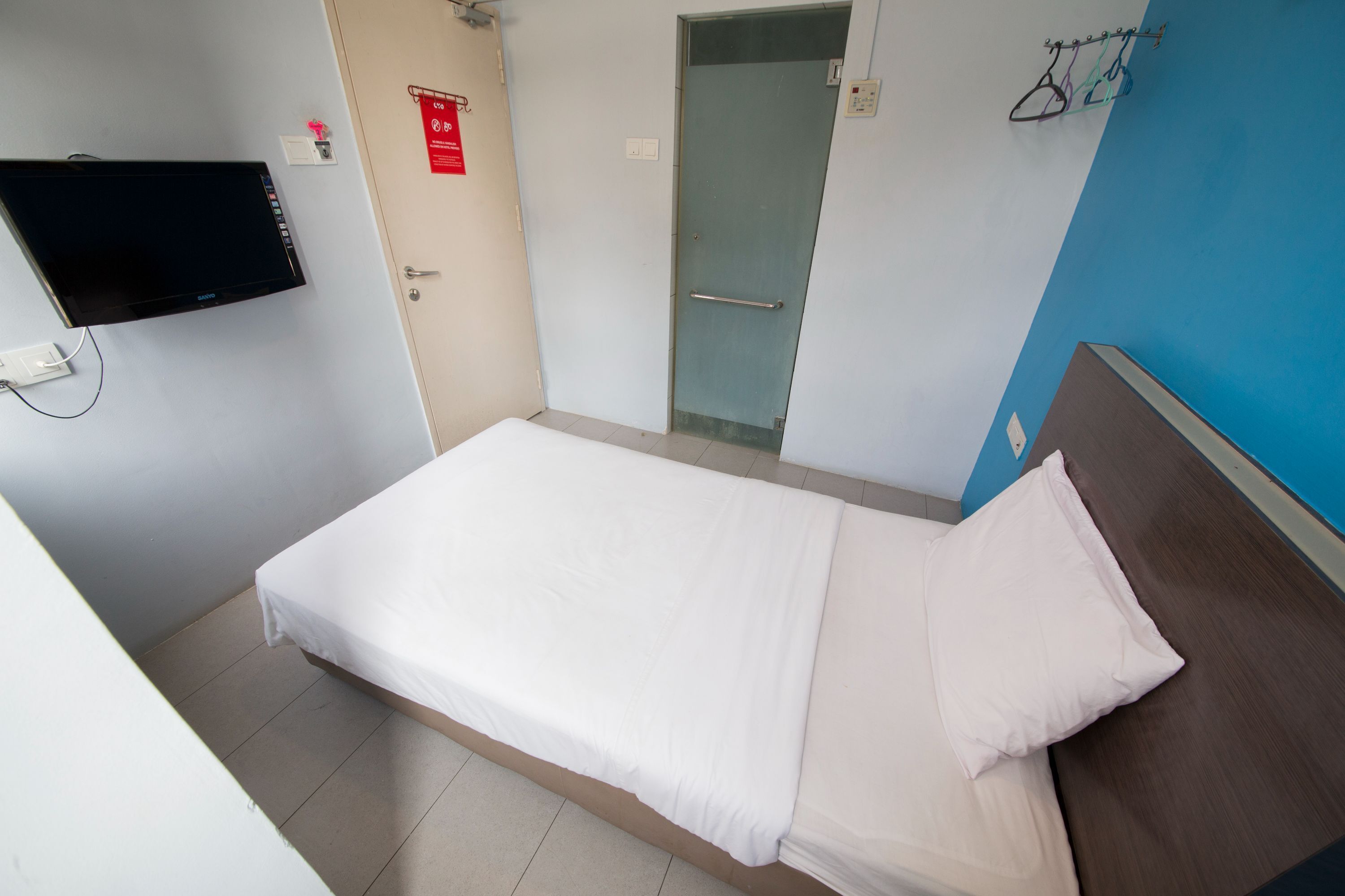 Una cama individual con ropa de cama blanca en una habitación con una pared azul decorativa, un televisor montado y una puerta con panel de vidrio.