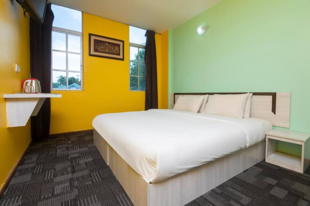 A hotel room with a bed, a nightstand, a window with curtains, and a wall with a framed picture.