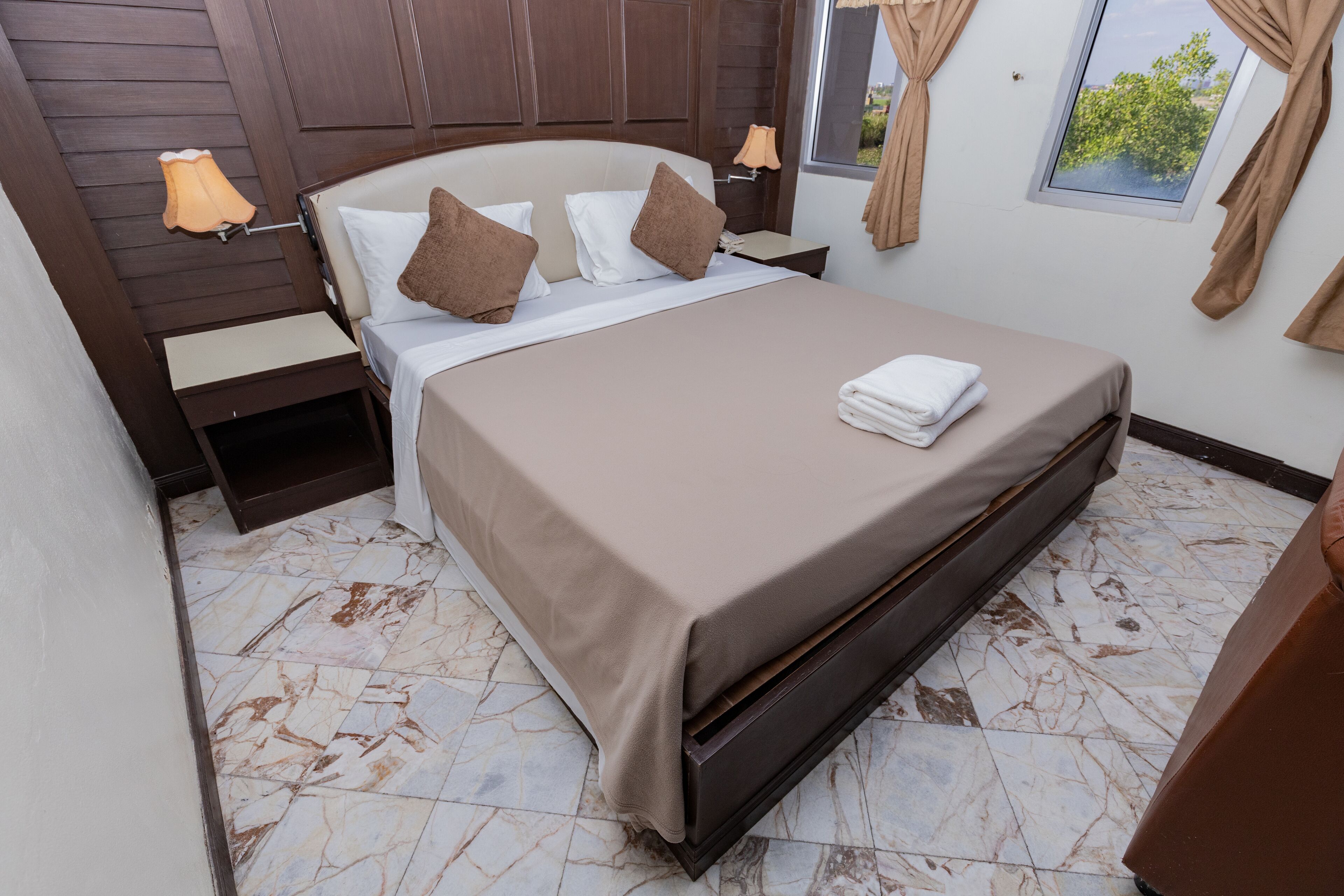 Un lit bien fait, recouvert d’une couvre-lit beige et agrémenté de deux oreillers, une table de chevet avec une lampe, et une fenêtre avec des rideaux.