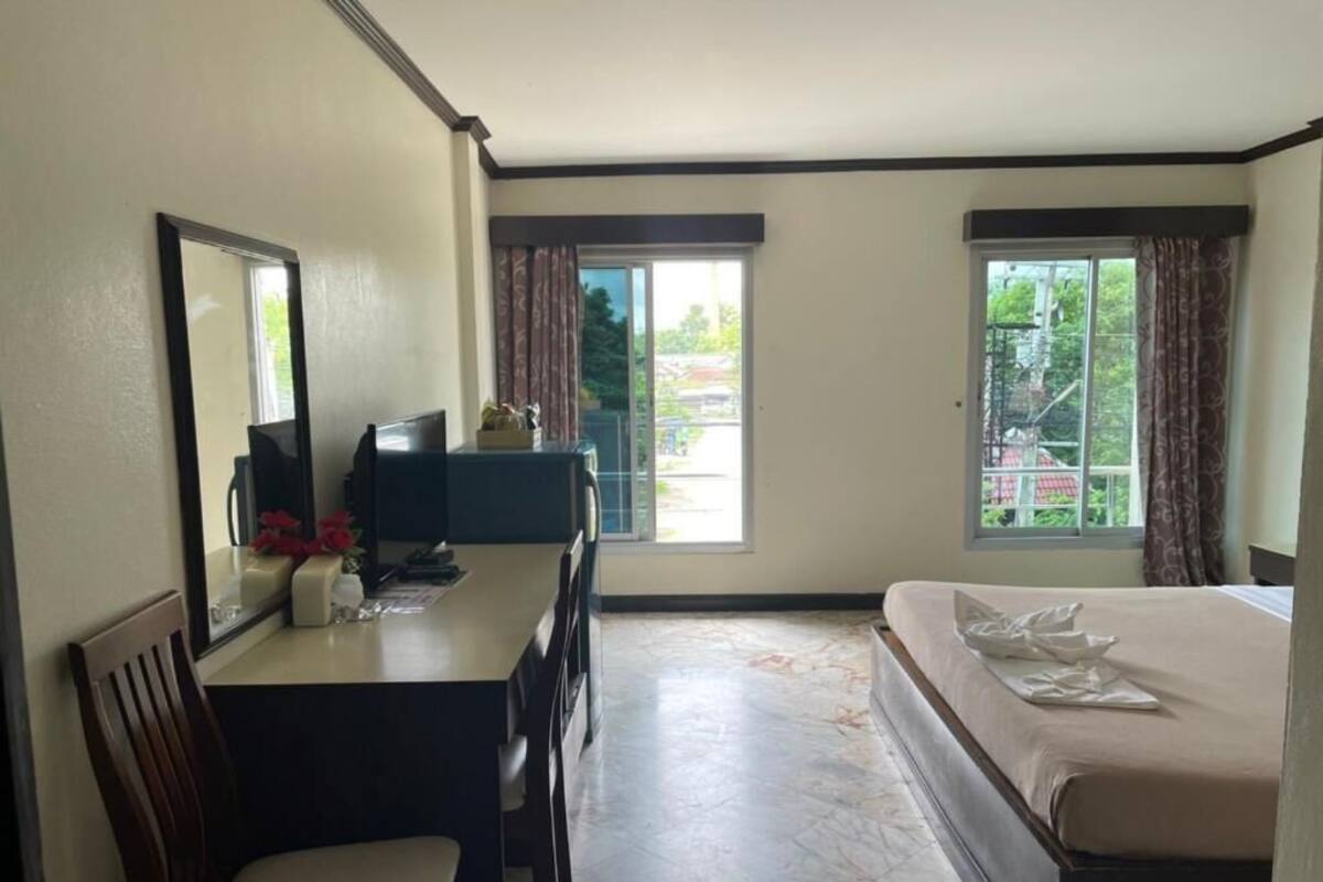 A hotel room with a bed, a desk with a chair, a mirror, and two windows with curtains.