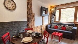 A cozy room with a round wooden table set for two, red chairs, a sofa with patterned cushions, a mounted television, and a rustic wooden pillar.