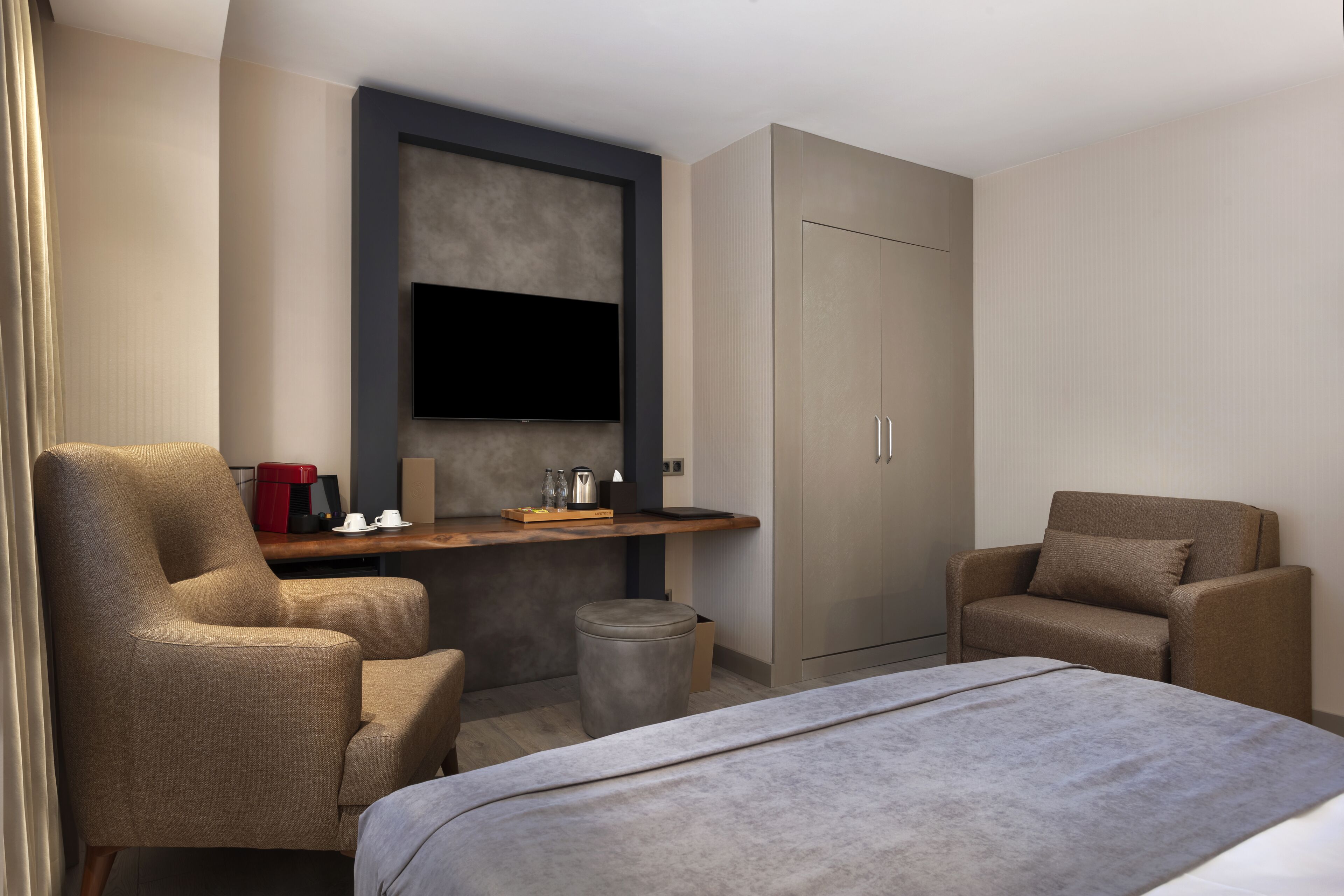 A modern hotel room with a bed, two armchairs, a small table with a coffee maker, and a wardrobe.
