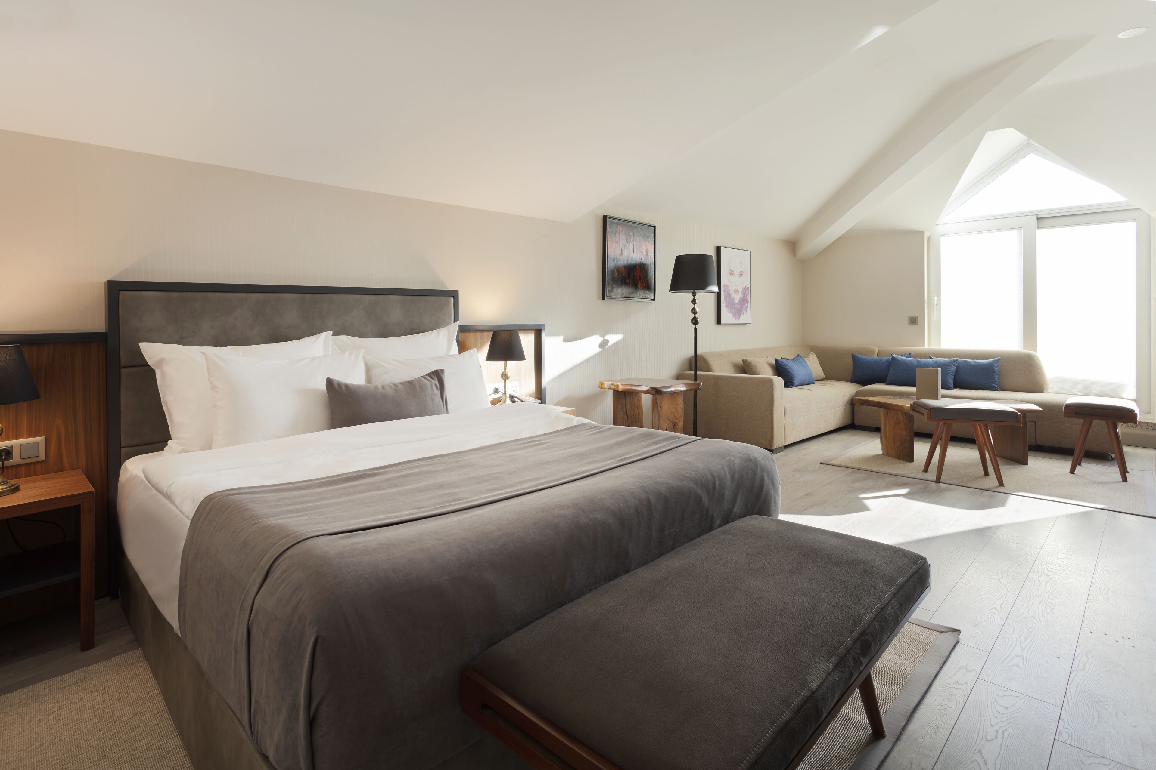 A modern hotel room with a large bed, a sofa, a desk, and a window providing natural light.