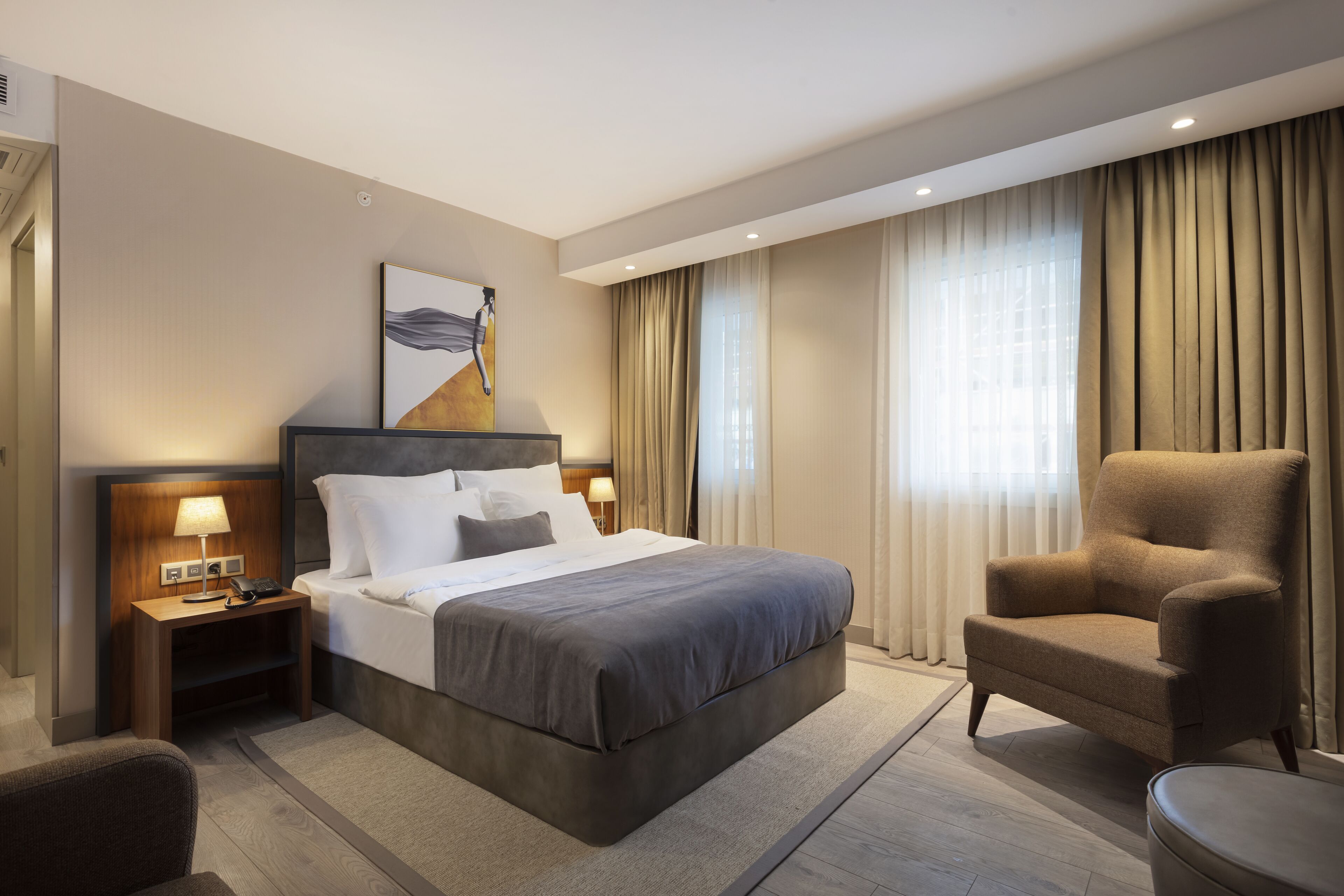 A modern hotel room with a large bed, a chair, a nightstand with a lamp, and a painting on the wall.