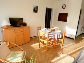 Un comedor con una mesa lista para dos personas, un televisor sobre un mueble de madera y un reloj en la pared.