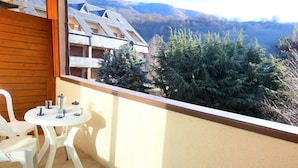 A balcony with a table and chairs, overlooking a mountainous landscape with coniferous trees and a building with a sloped roof.