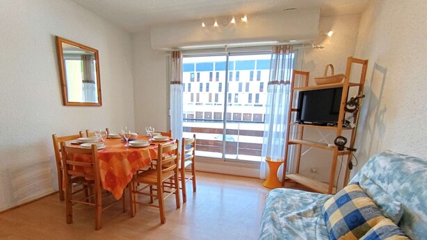 Un coin repas avec une table dressée pour quatre personnes, un canapé, un téléviseur posé sur un meuble et un balcon offrant une vue sur les bâtiments.