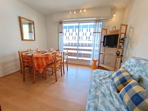 A dining area with a table set for four, a sofa, a TV on a stand, and a balcony with a view of buildings.