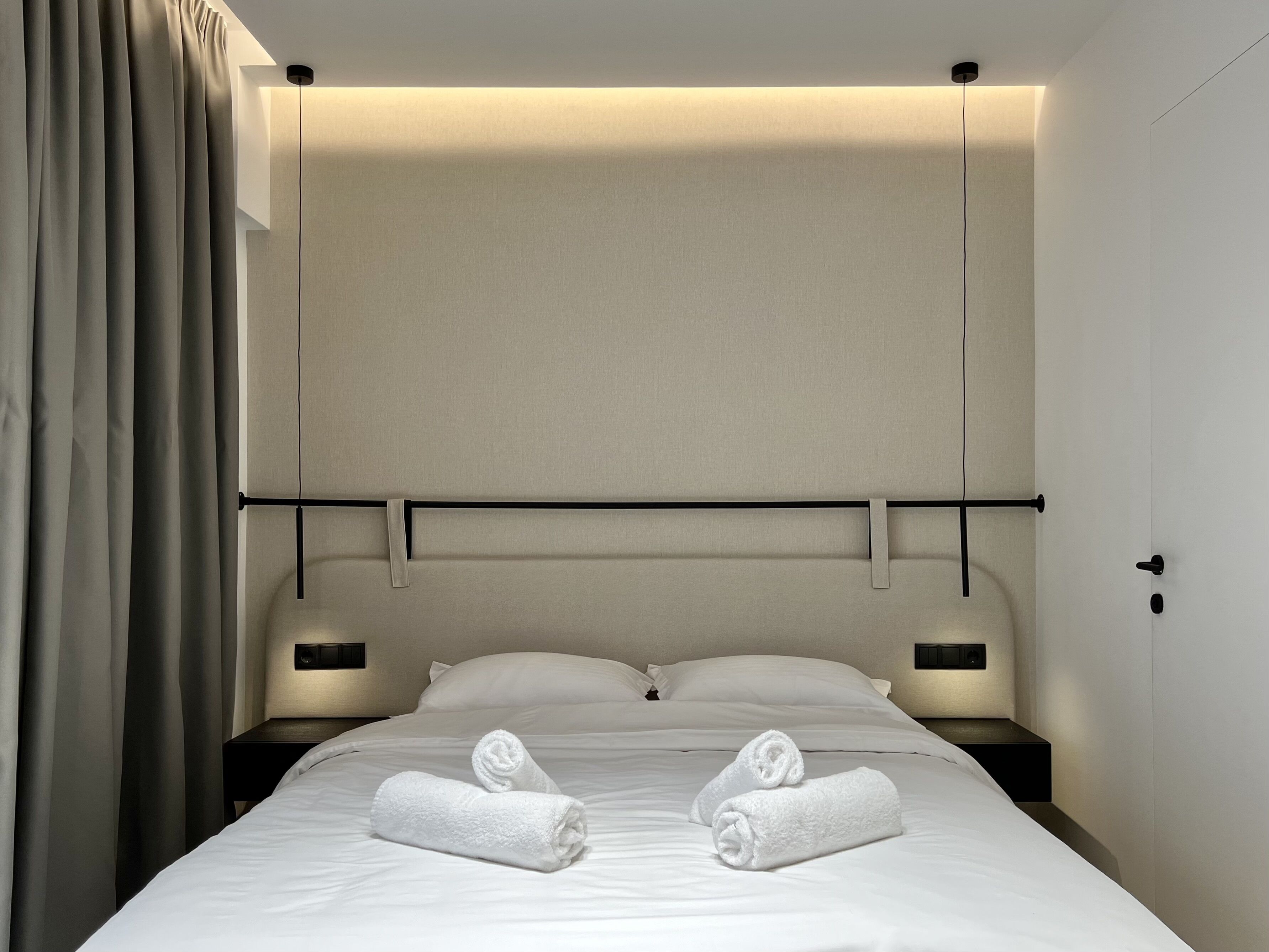 A hotel room with a neatly made bed, two rolled towels, a headboard, bedside tables, and a curtain.
