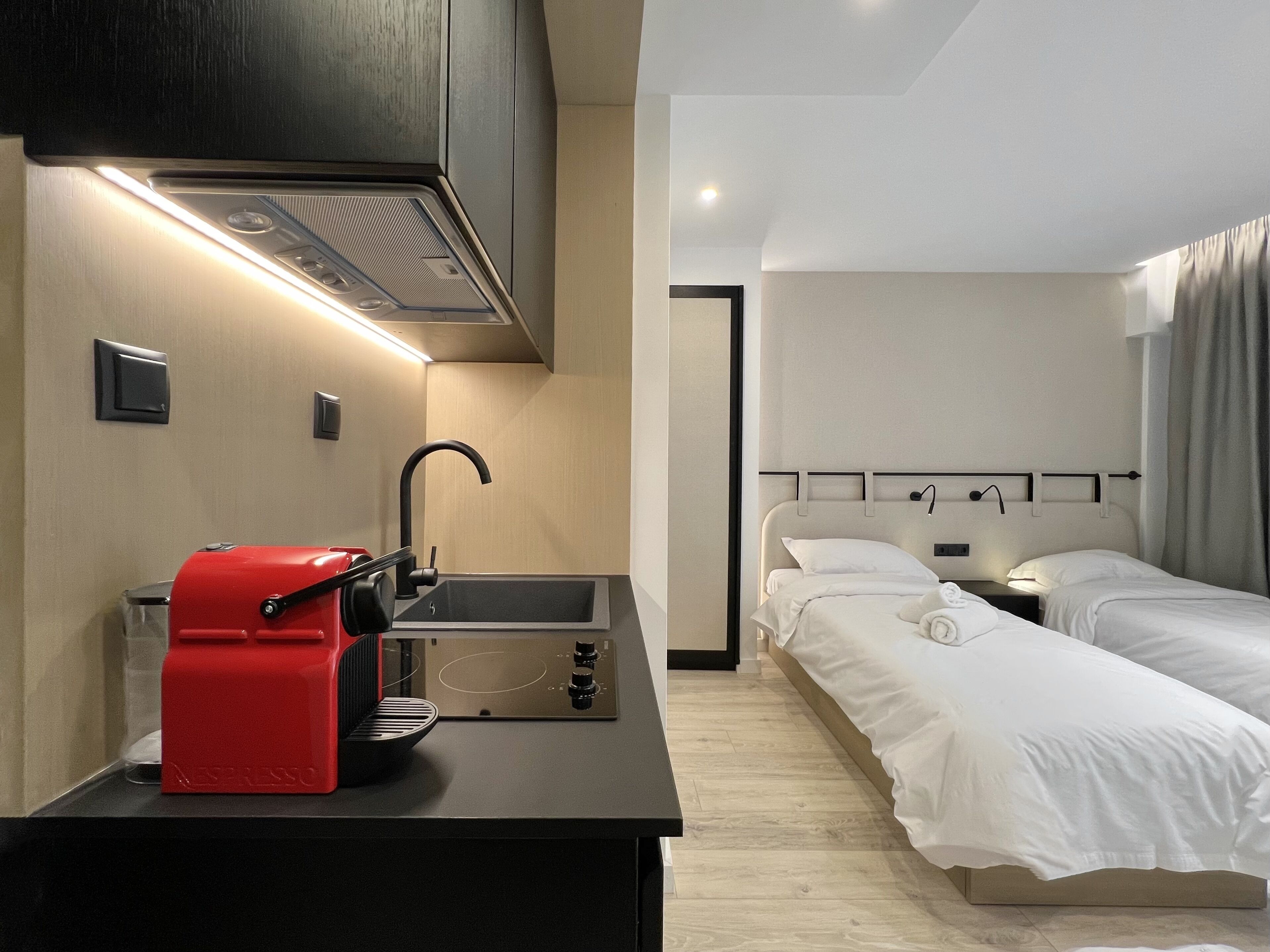 A modern hotel room with a kitchenette, a bed with two pillows, a bedside table, and a nightstand.