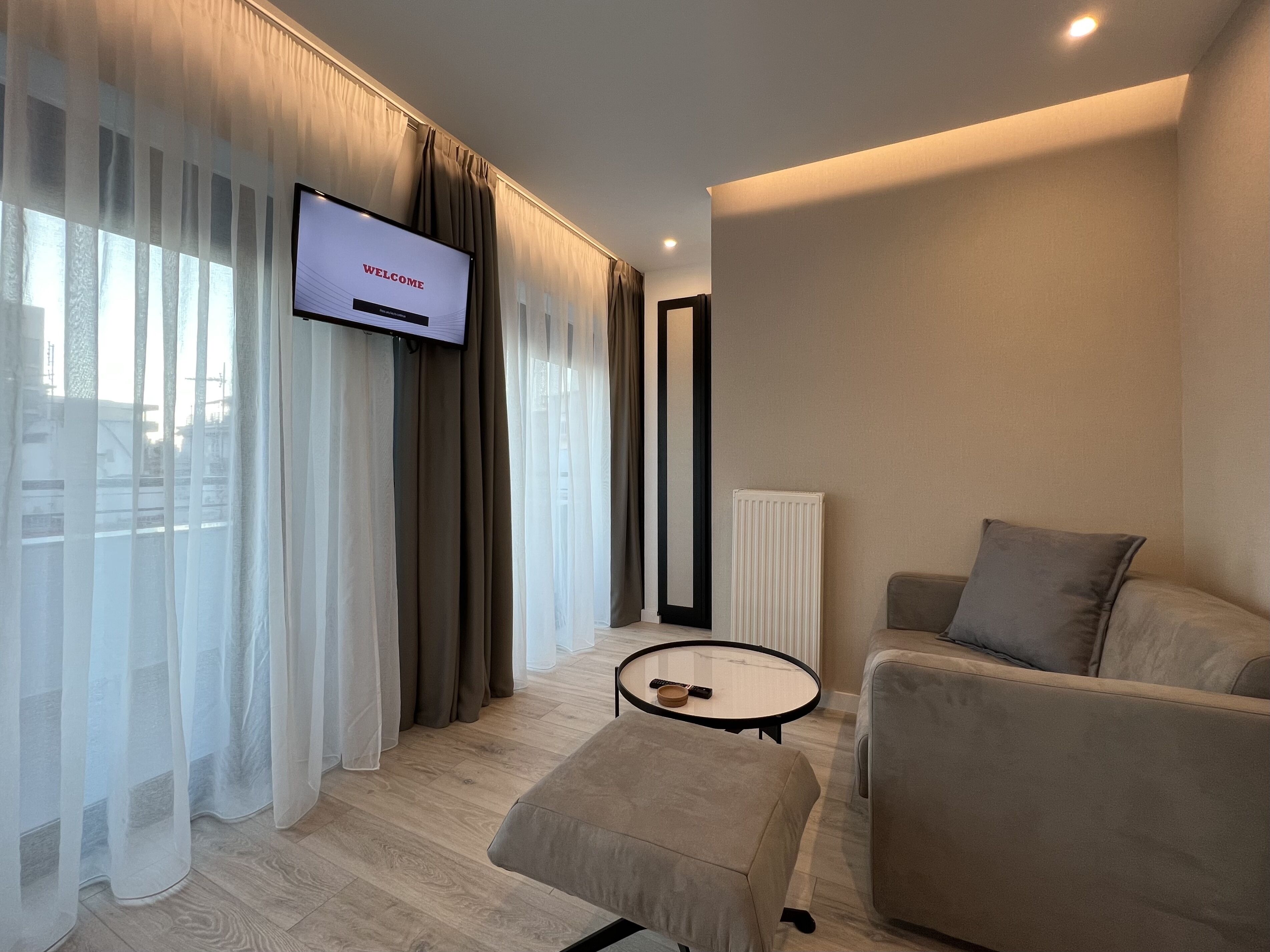 A modern living room with a flat-screen TV displaying "WELCOME", a sofa, a coffee table, and sheer curtains.