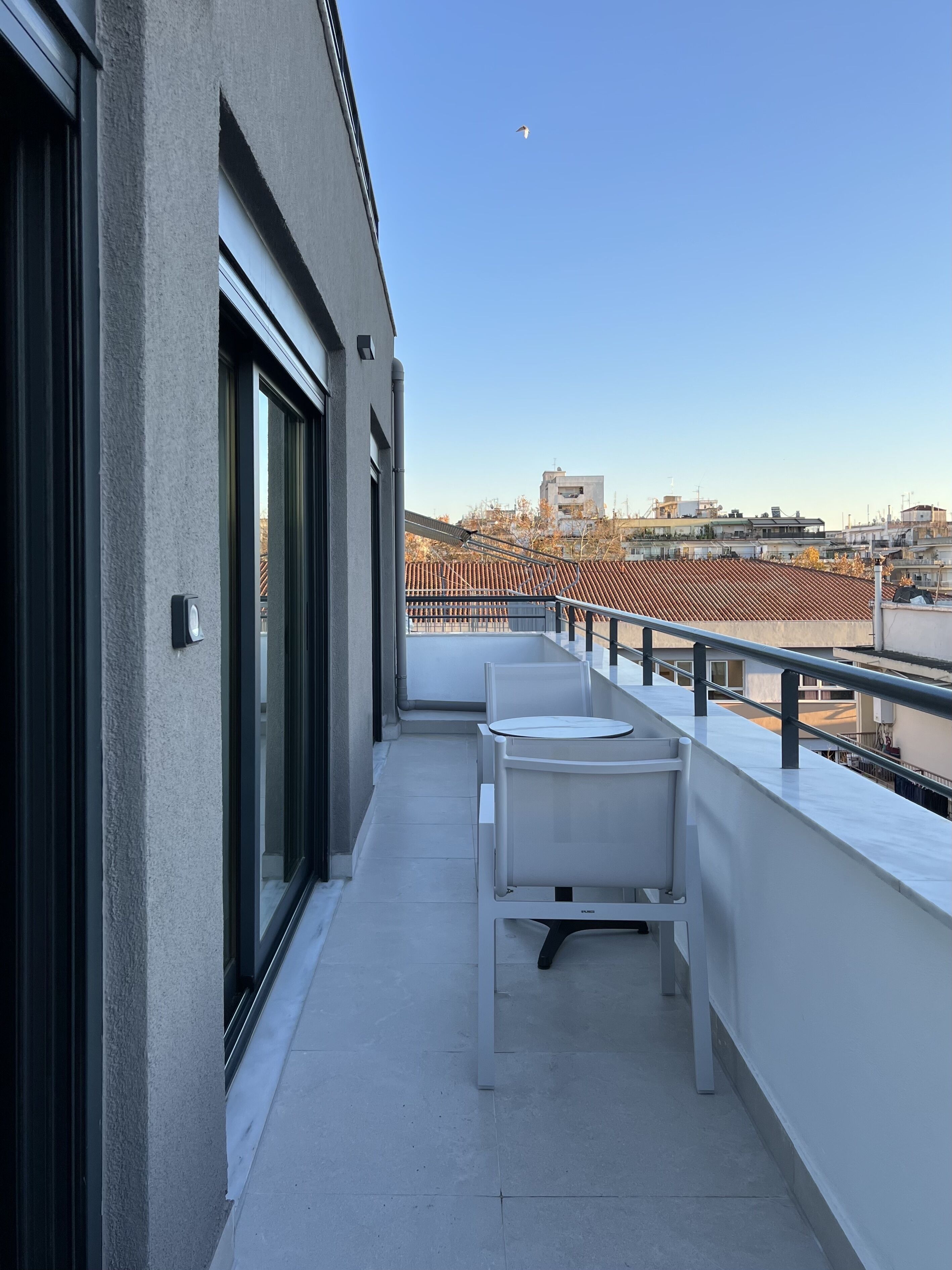 A balcony with two white chairs and a table, overlooking a cityscape with buildings and a clear sky.