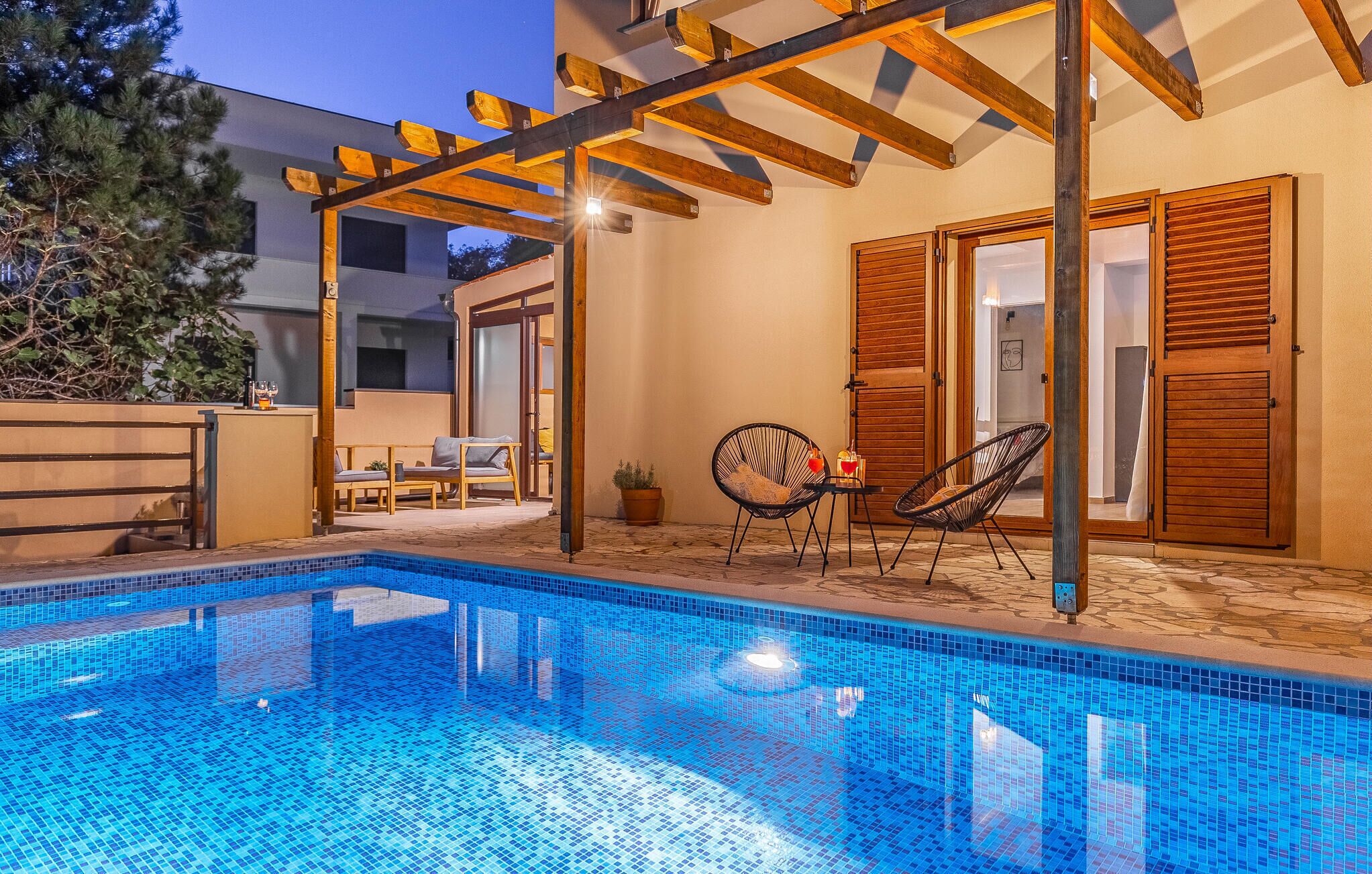 En pool med ett kaklat poolområde, en träpergola och två solstolar.