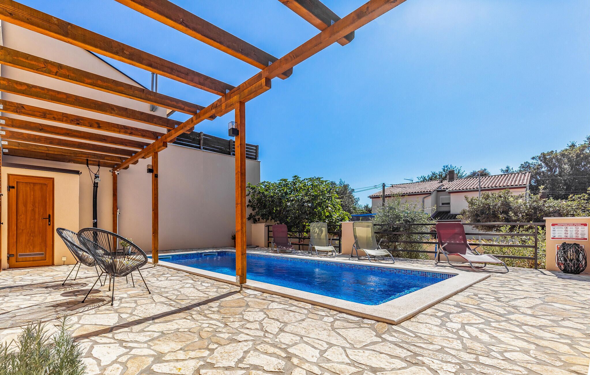 A pool area with stone flooring, wooden pergola, and lounge chairs.
