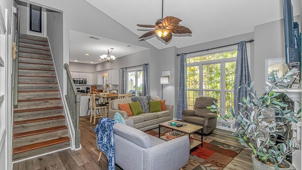 A spacious living room with a grey sofa, a coffee table, and a ceiling fan. A staircase leads to a kitchen area with white cabinets and a dining table. - Discount! Walk to Beach, Golf,  Pool, Arcade, Beautiful and peaceful community (North Myrtle Beach)