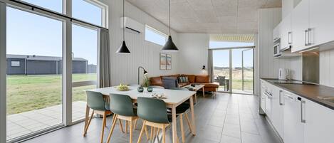 A modern kitchen with white cabinets, a dining area with a table and chairs, and large windows offering a view of the outdoors.
