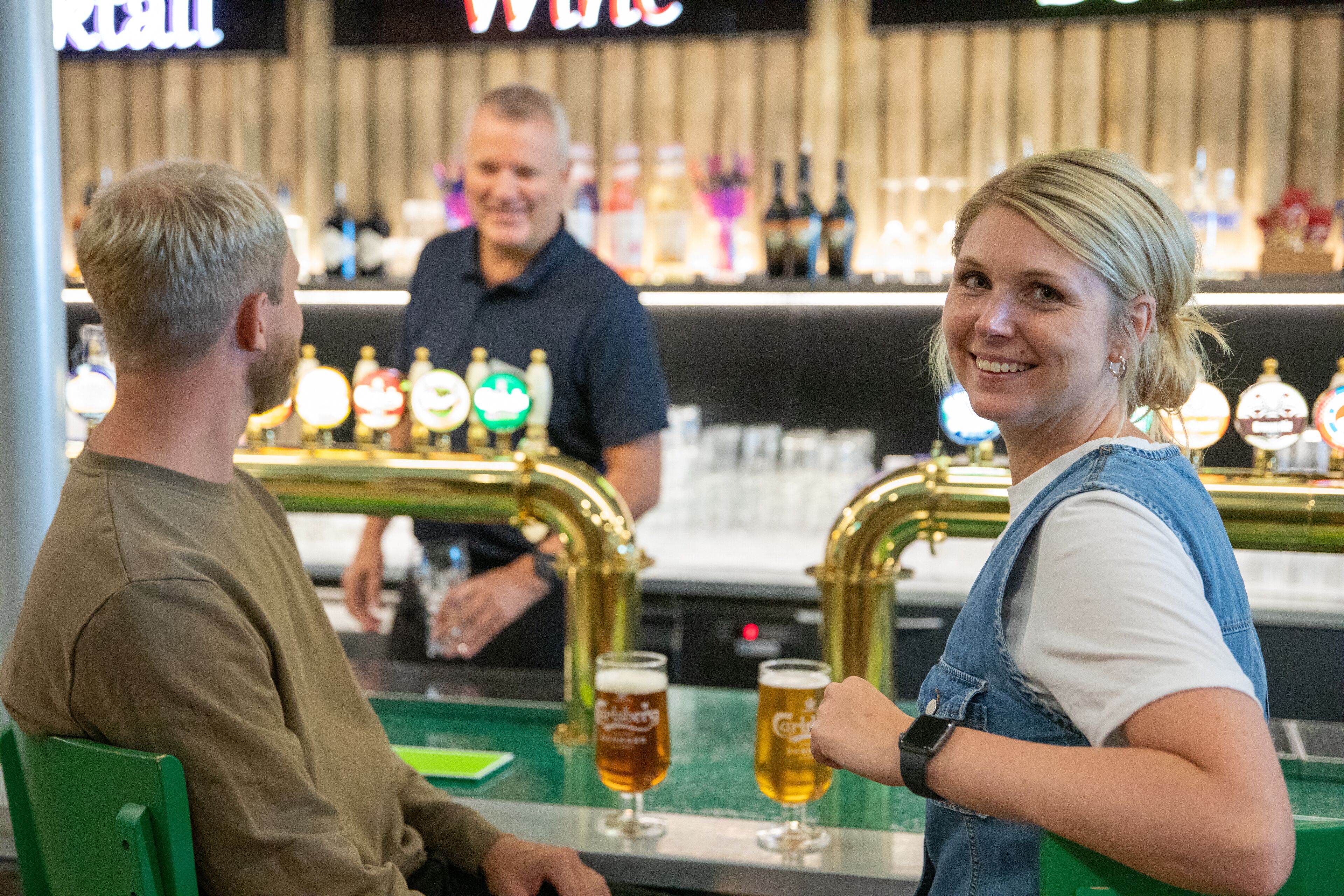 En bar med två personer sittande vid ett bord, en bartender i bakgrunden och ett urval av ölkranar.