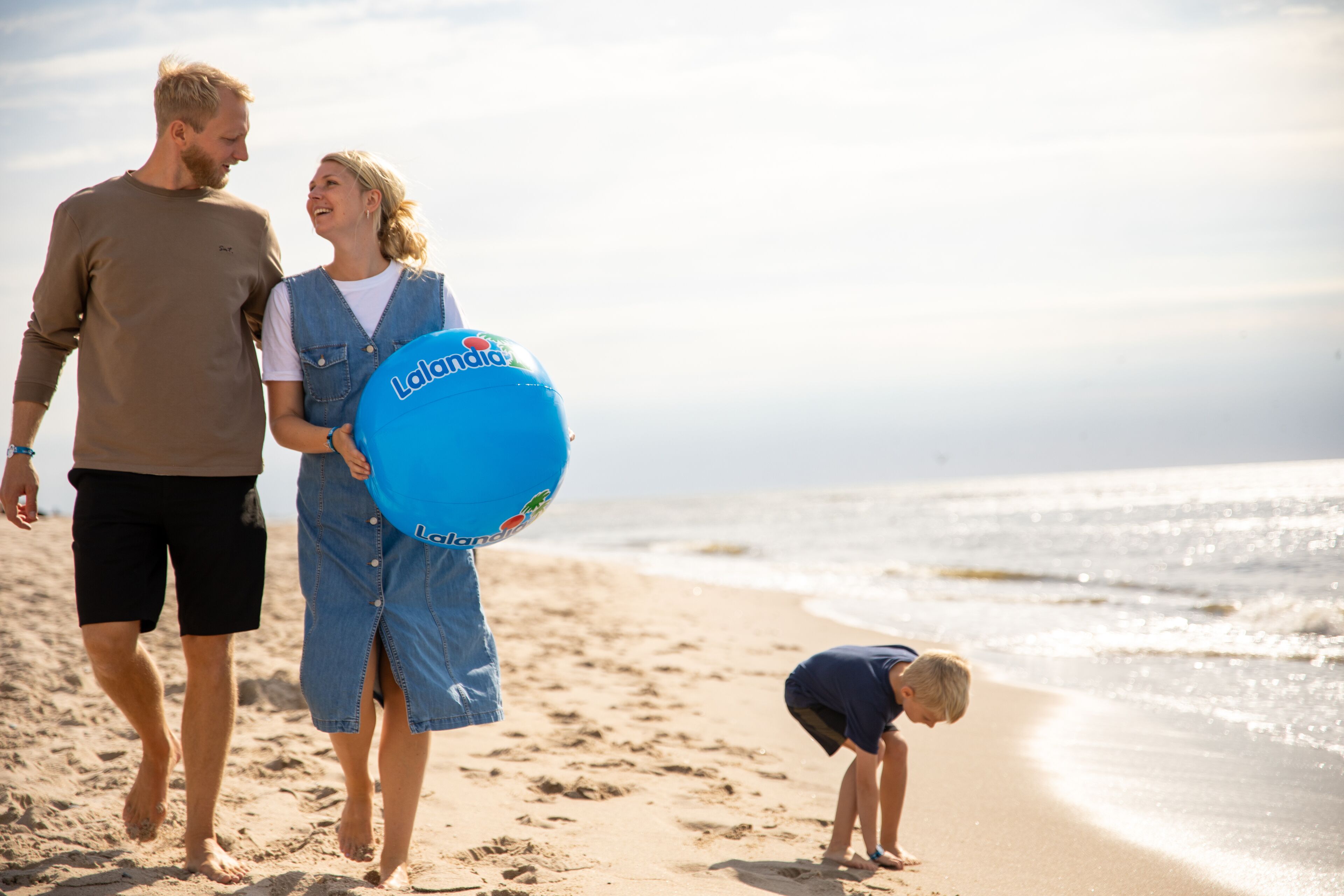 Escena de playa con una familia caminando por la orilla, un niño jugando en la arena y un flotador inflable azul.