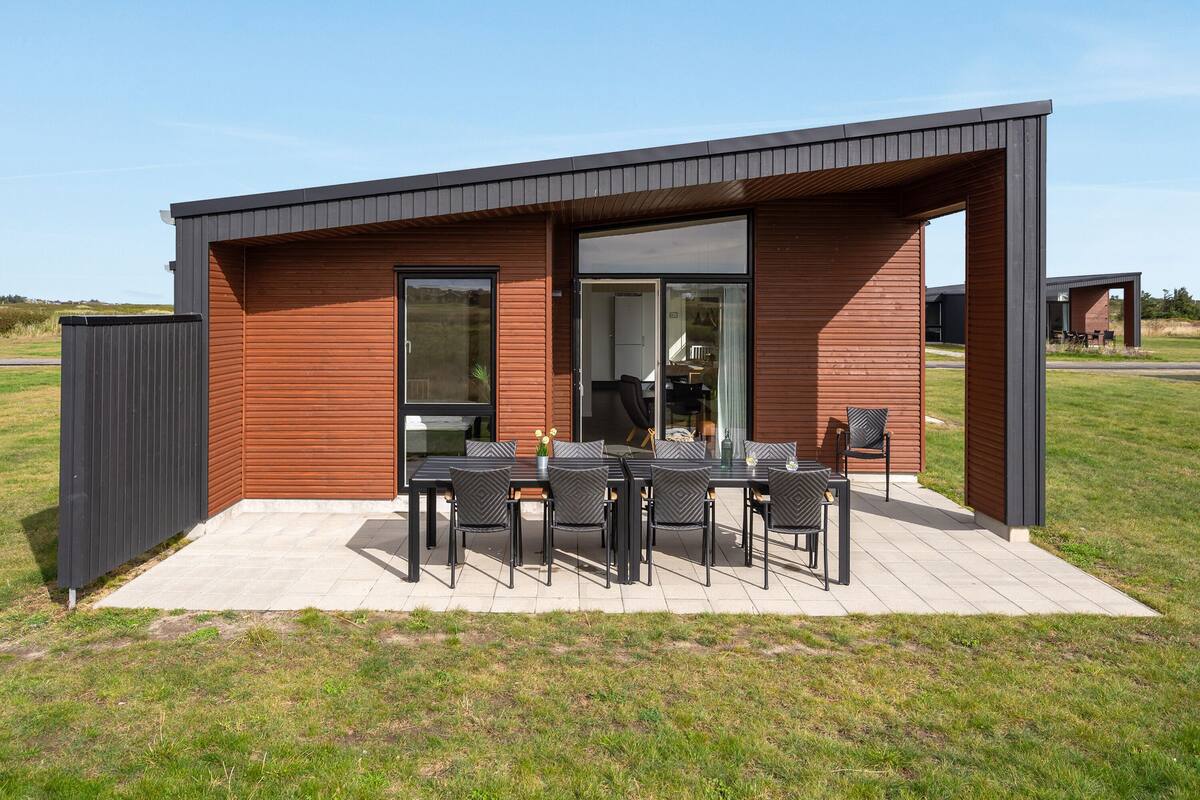 Un área de comedor exterior moderna con fachada de madera, una mesa de patio con sillas y un ventanal de vidrio que da a un espacio interior.