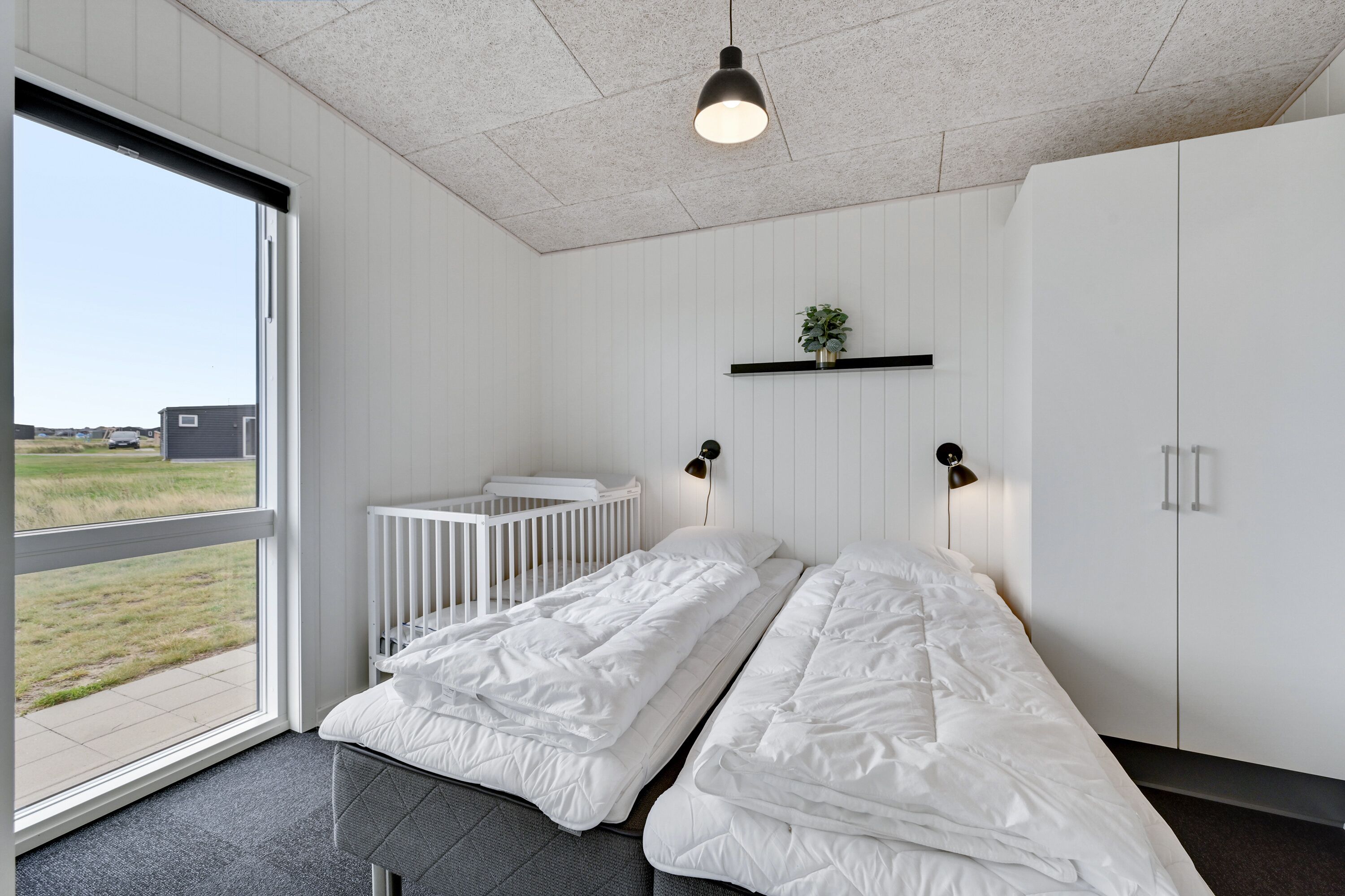 Une chambre avec une grande fenêtre, un berceau, un lit avec une literie blanche, une plante posée sur une étagère et une armoire.