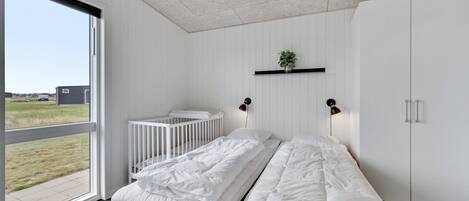 Une chambre avec une grande fenêtre, un berceau, un lit avec une literie blanche, une plante posée sur une étagère et une armoire.
