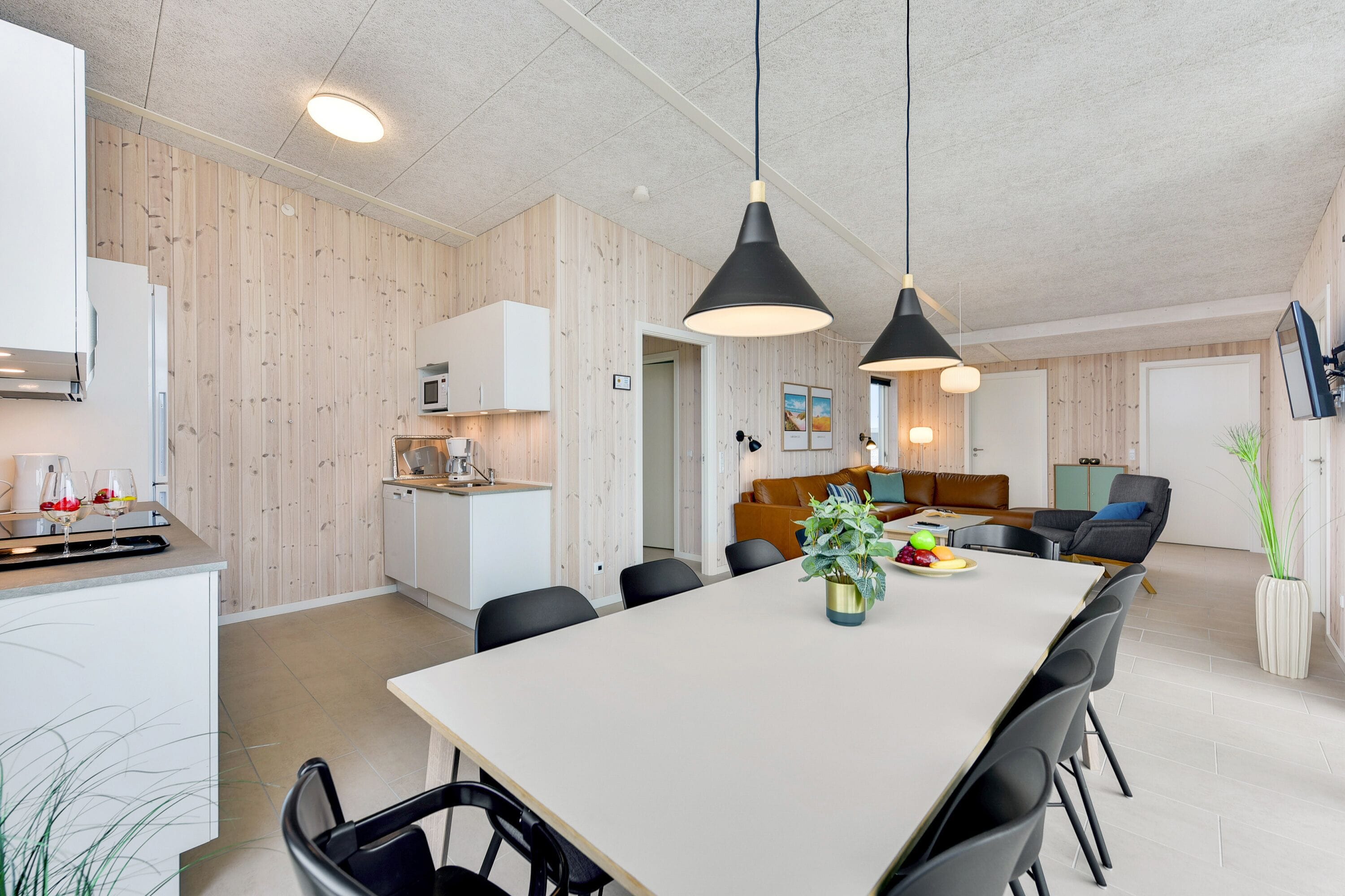Eine moderne Küche mit Holzvertäfelung, ein Essbereich mit großem Tisch und Stühlen sowie ein Wohnbereich mit Sofa und Couchtisch.