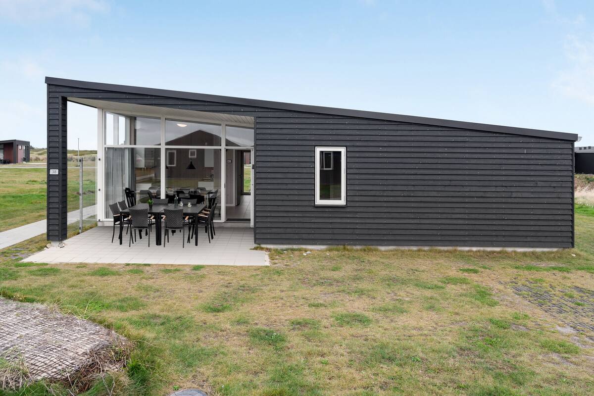 Un edificio moderno de una sola planta con fachada oscura, grandes puertas correderas de vidrio y un patio exterior con mesa y sillas.