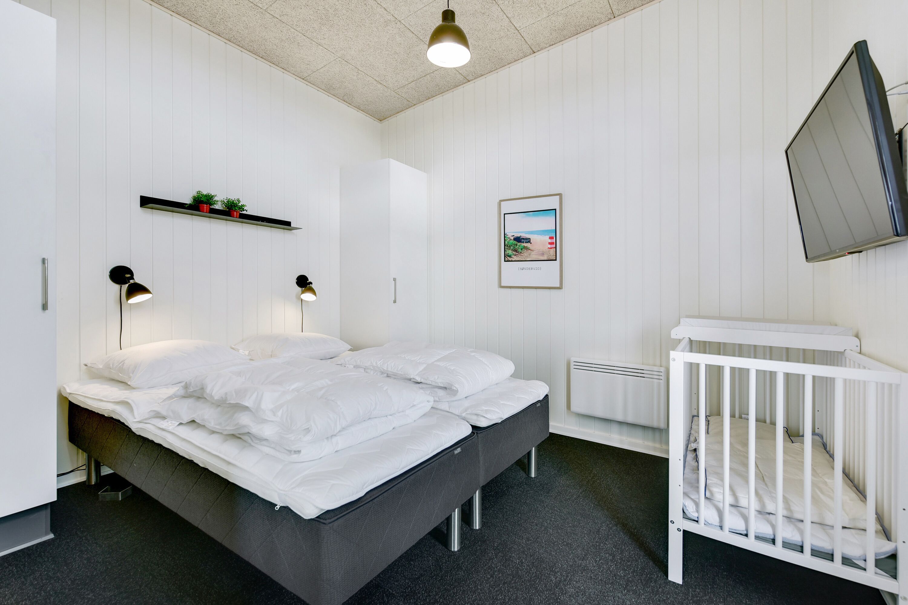 A modern bedroom with a bed, bedside lamps, a crib, a shelf with plants, and a framed picture on the wall.
