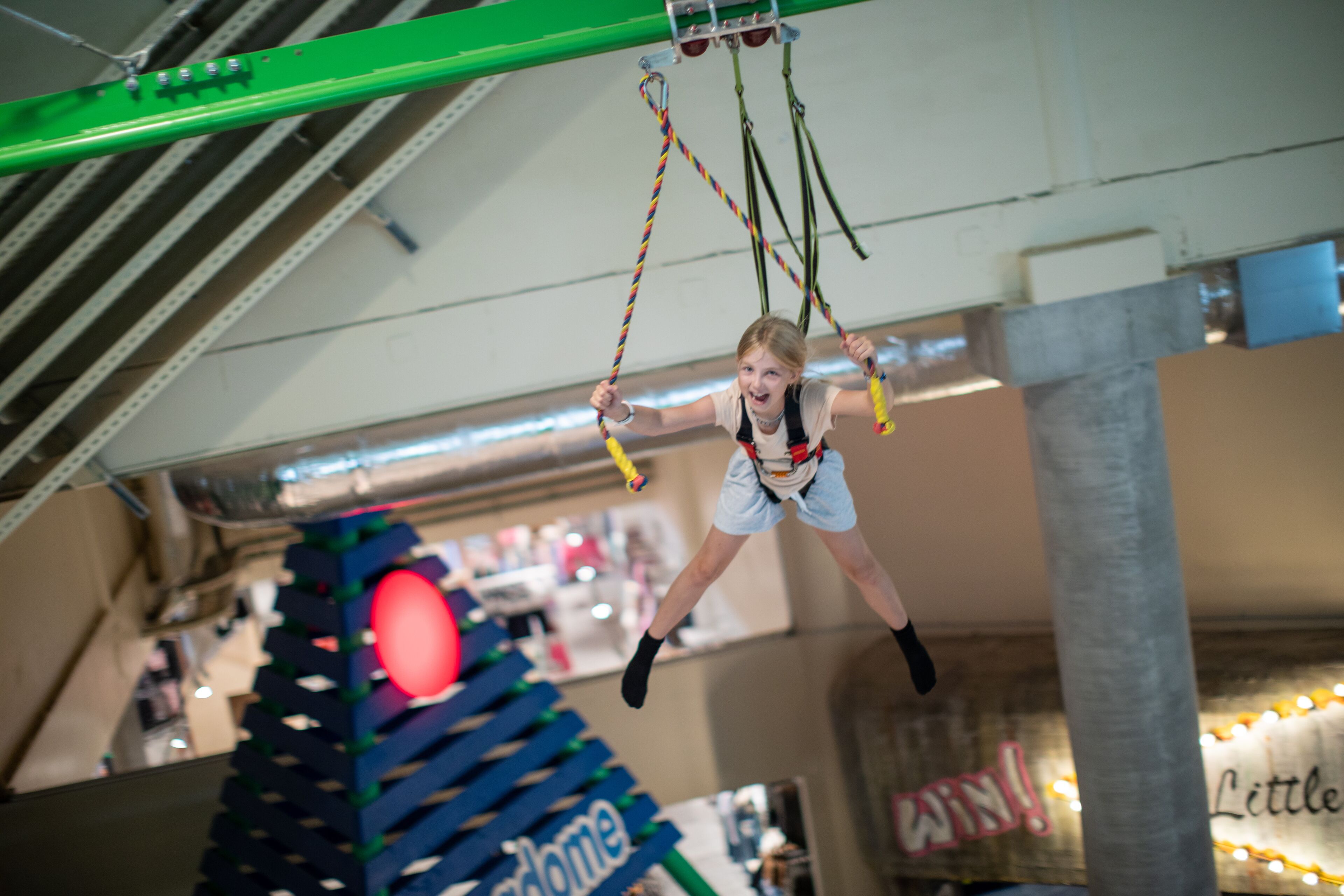 A person swinging on a rope in an indoor setting with visible structural elements and signage.