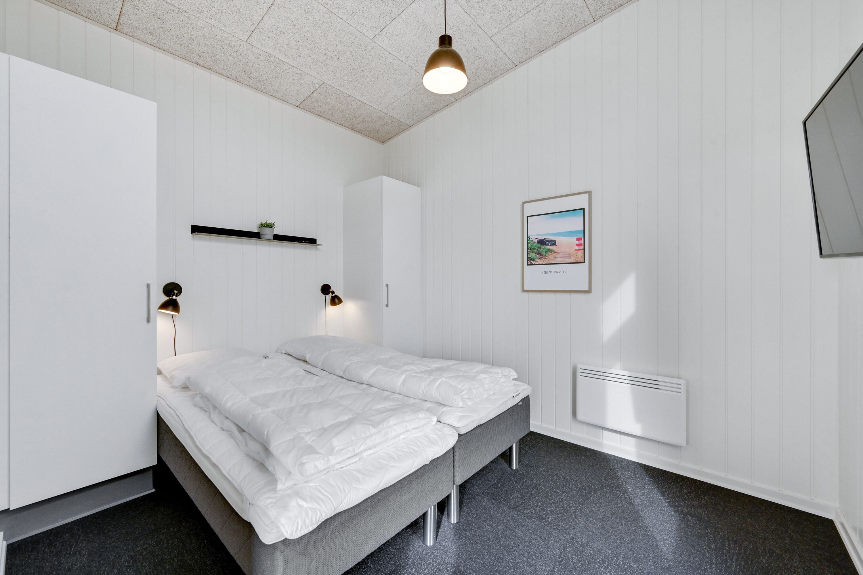 Un dormitorio con una cama, lámparas de noche, una estantería con una planta, un cuadro enmarcado y un radiador.
