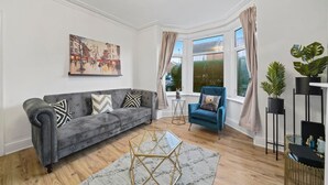 Una sala de estar con un sofá gris, un sillón azul, una mesita de centro de cristal y un cuadro en la pared.