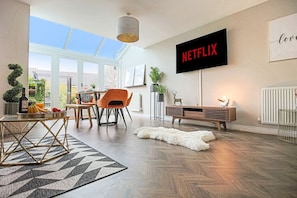 Ein modernes Wohnzimmer mit einem großen Fernseher, der 'NETFLIX' anzeigt, einem Essbereich mit Tisch und Stühlen sowie einem Blick ins Freie durch große Fenster.