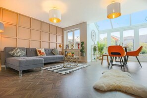 Ein modernes Wohnzimmer mit grauem Sofa, orangenen Sesseln, einem Holztisch und einem großen Fenster mit Ausblick.