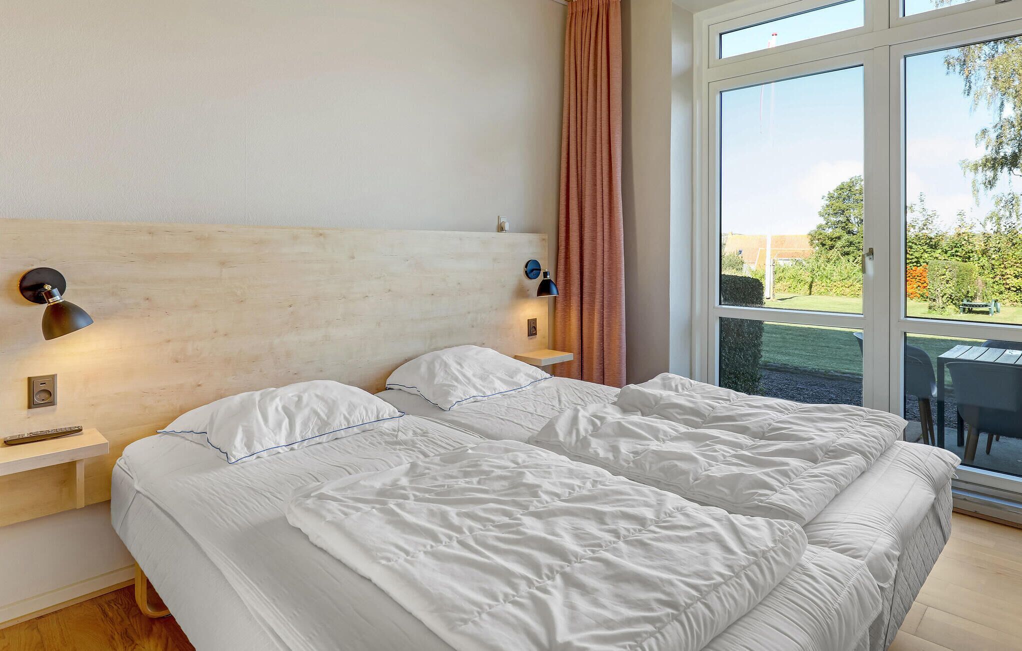 A hotel room with a large bed, two bedside lamps, a wooden headboard, and a view of the outdoors through a large window.