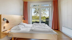 Ein Schlafzimmer mit einem hölzernen Kopfteil, einem Bett mit weißer Bettwäsche, einem Nachttisch, einer Lampe und Blick ins Freie durch ein großes Fenster.
