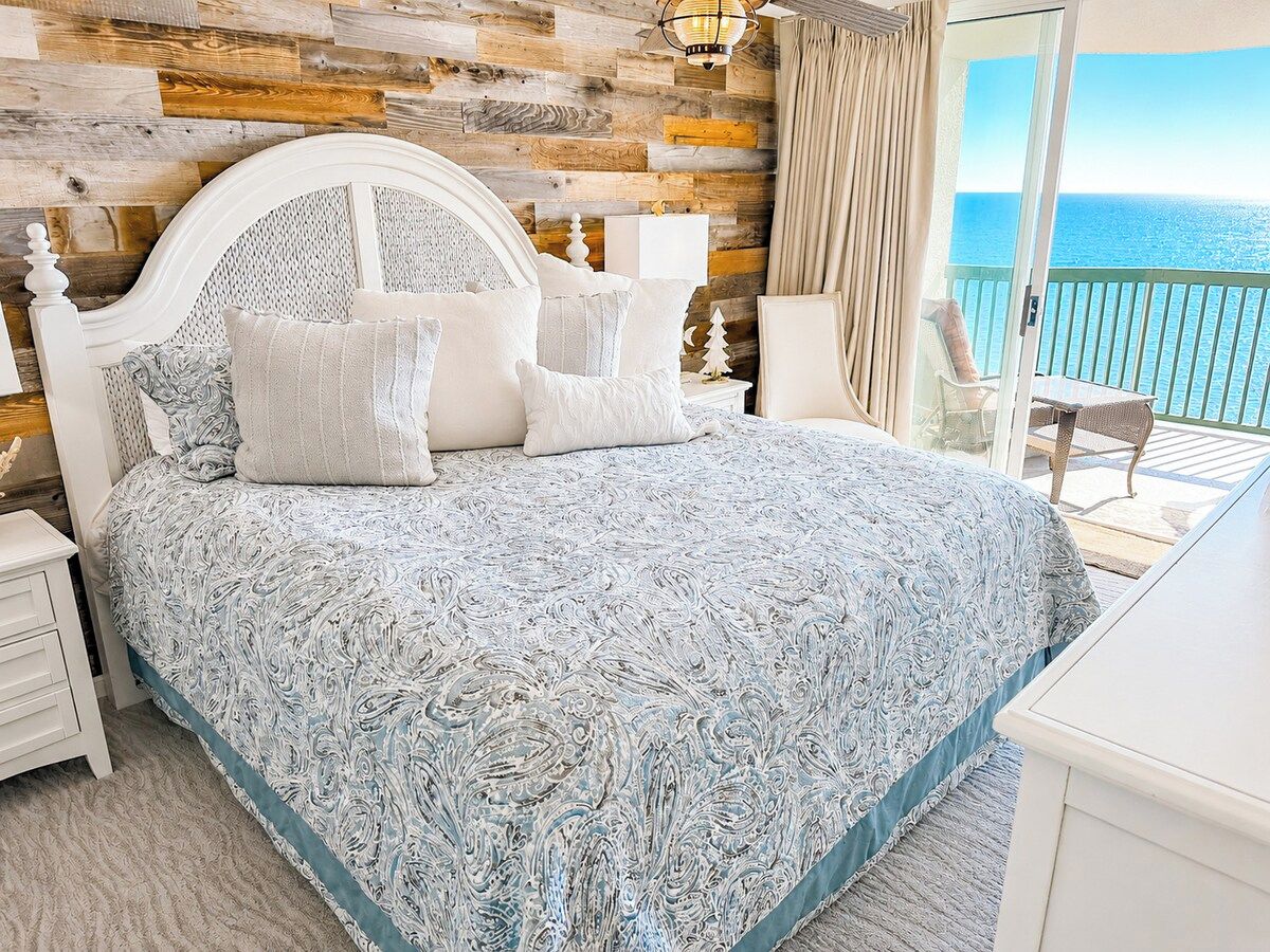 A bedroom with a bed, nightstands, a dresser, and a view of the ocean.