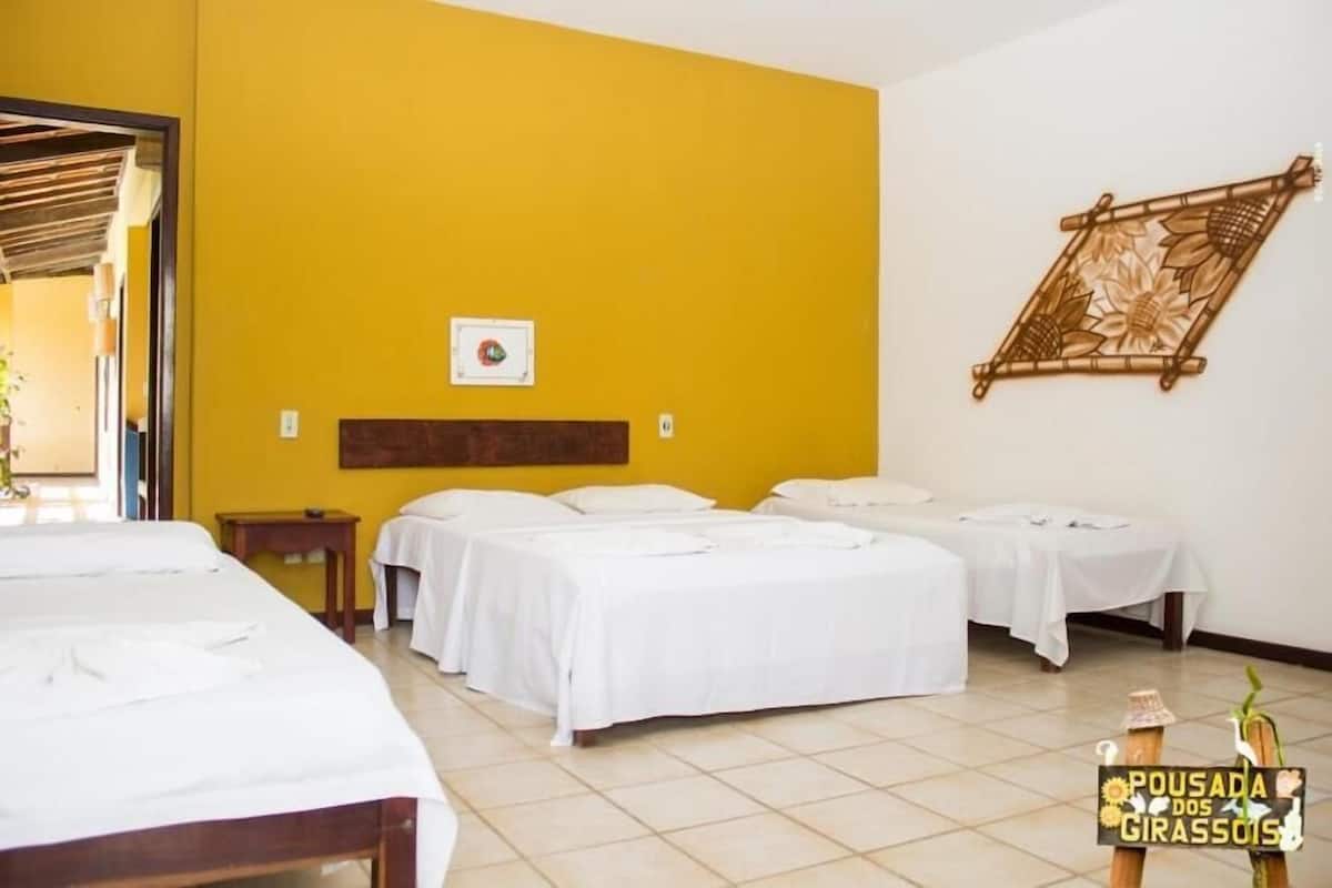 A hotel room with three beds, a wooden headboard, a wall-mounted decorative piece, and a small table with a plant.