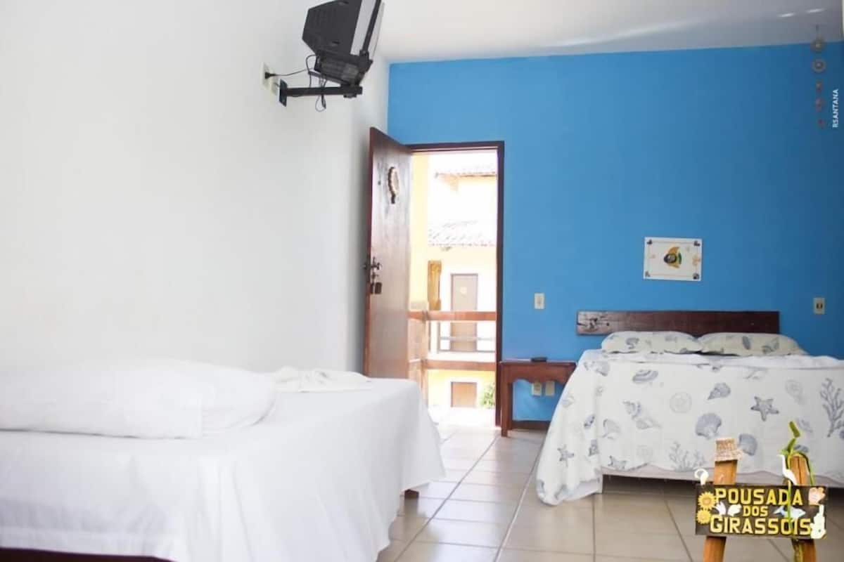 A room with two beds, one with a white bedspread and the other with a patterned one. There is a blue wall and a door leading to an outdoor area.