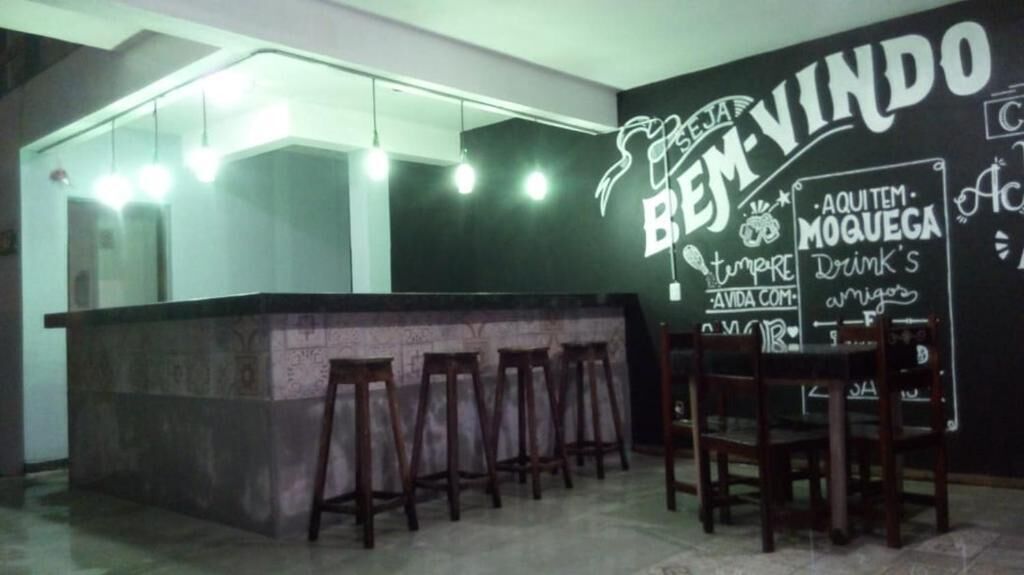 Un espace bar avec un tableau noir sur lequel on peut lire les inscriptions « BEN-VINDO » et « AQUITEM MOQUEGA Drink’s ». On y trouve des tabourets en bois et un comptoir de bar.