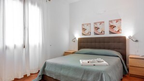 Un dormitorio con cama, mesita de noche, cortinas en la ventana y una obra de arte en la pared.