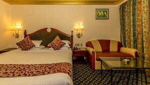 Habitación de hotel con una cama grande, dos lámparas de noche, un sofá, una mesa de centro de vidrio, alfombra estampada y un cuadro en la pared.